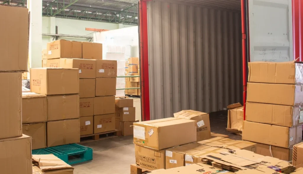 倉庫の荷役ドックにおいて、開いたコンテナの近くに積み上げられた段ボール箱とパレット。