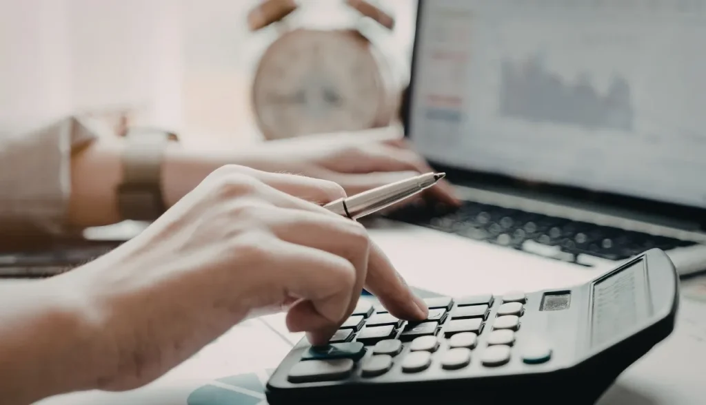 Close-up of hands using a calculator and pen, with a laptop displaying data charts in the background.