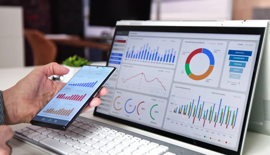 Person viewing real-time inventory analytics dashboards on a smartphone and laptop, showing charts and graphs that RFID-powered inventory systems provide.