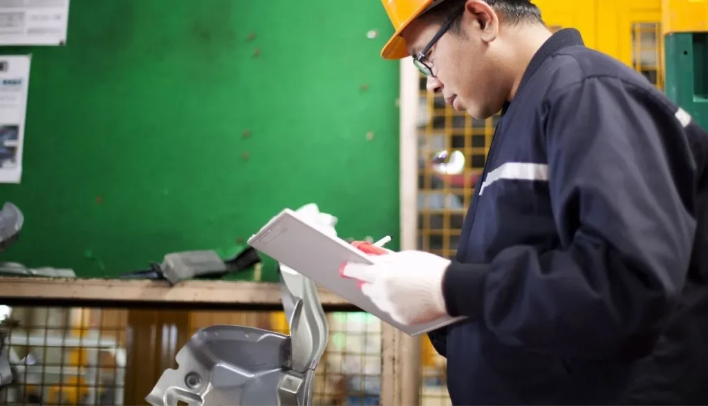 Factory inspector wearing safety gear writes records on a clipboard while checking manufactured parts on the production floor.