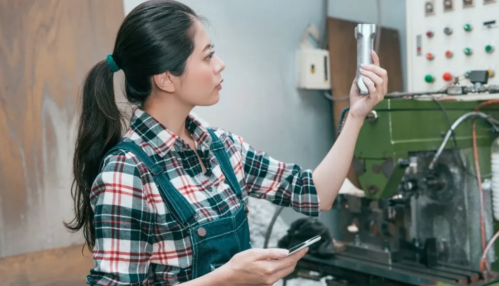 Female factory worker inspects a metal workpiece while referencing data on a mobile smartphone on the production line.